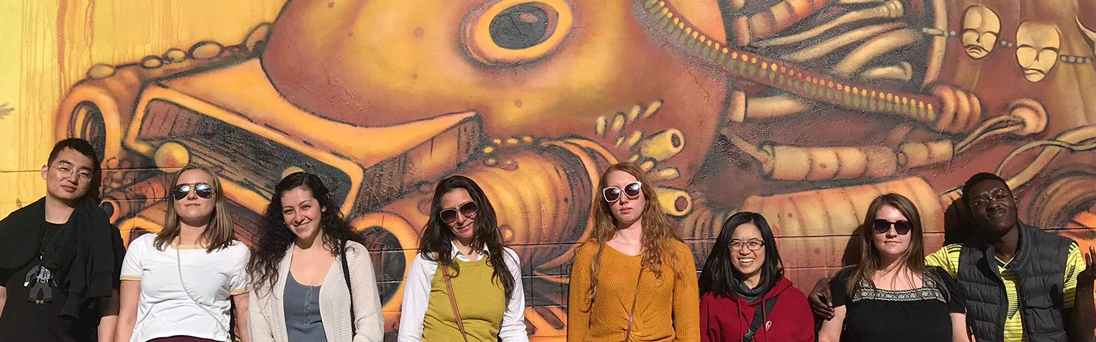 Students are in a line against a wall with a piece of street art on it. some students are smiling and some have straight-faces.